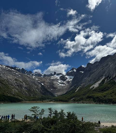       Wide valley surrounded by dramatic craggy peaks with patches of snow beneath a bright blue, cloud-dotted sky.
  