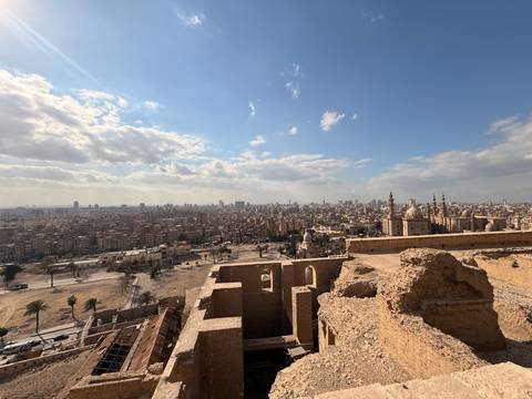       Panoramic view over Cairo with historic mosque domes and minarets in the foreground under a bright sky.
  