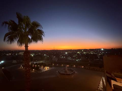       Cityscape at dusk with palm tree silhouette and vibrant orange horizon transitioning to deep blue night sky.
  