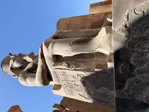       Massive seated statue of Ramesses II carved from sandstone against a clear blue sky.
  