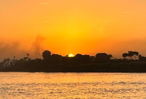       An intense orange sunset over the Nile river, silhouetting palm trees and low buildings with smoke rising in the distance.
  