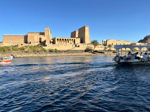       A boat of tourists approaches Philae Temple, whose ancient sandstone walls rise beside the Nile under a clear blue sky.
  