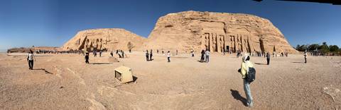       A wide panorama of Abu Simbel with crowds exploring the colossal rock-cut statues under a cloudless sky.
  