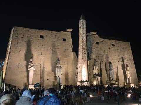       Luxor Temple entrance and obelisk illuminated at night, crowds at the base.
  