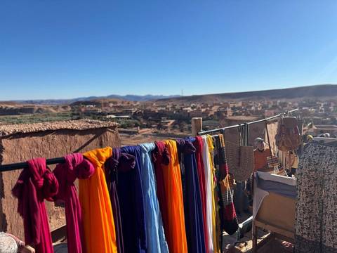       Vivid fabric scarves hang drying above a desert village with distant ranges under blue sky.
  