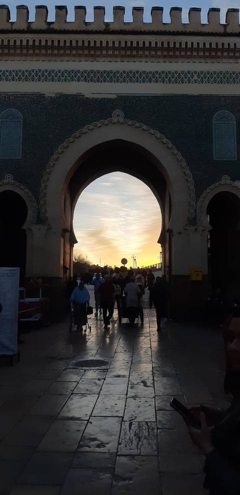       Crowd silhouettes pass through a monumental Moroccan gate at sunset with orange sky.
  