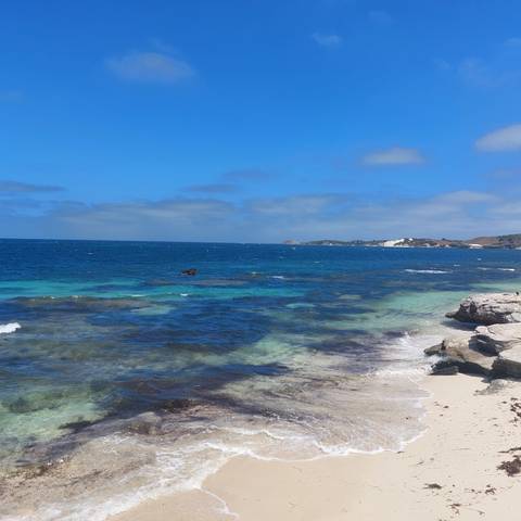       Clear turquoise ocean laps rocky shoreline under bright blue sky with distant sandy headland.
  