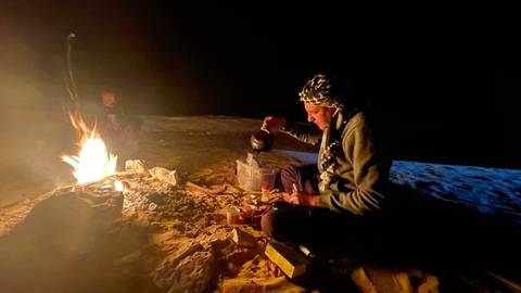       Man in keffiyeh pours tea beside a small camp-fire on desert sand at night
  