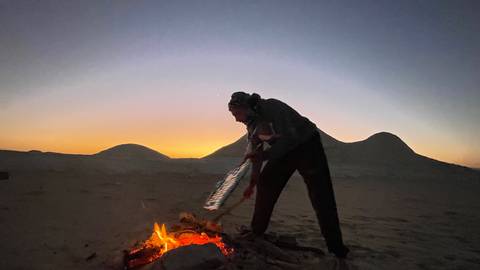       Silhouette of a guide tending a desert fire at dawn against pastel horizon and dunes
  