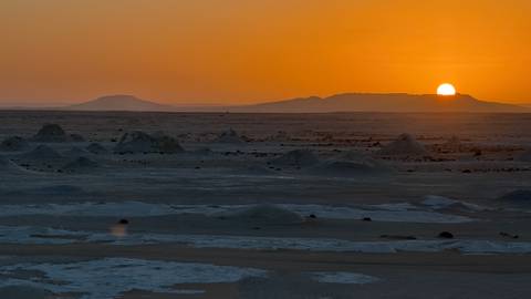       Orange sun sets over vast flat desert peppered with chalk mounds
  