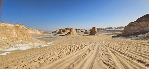       Tracks in golden sand lead toward clustered desert pinnacles beneath clear blue sky
  