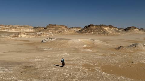       Single traveller dwarfed by expansive sandy plain and low rocky hills
  