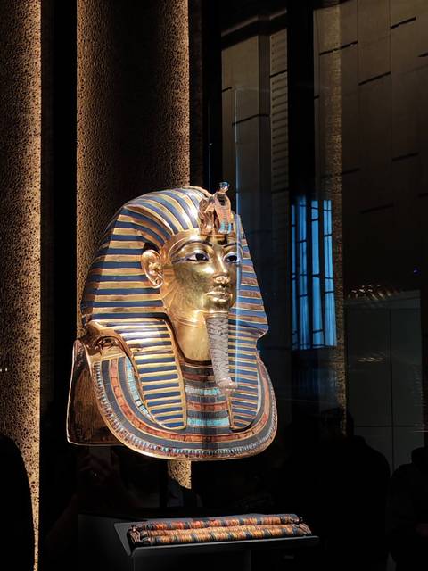       Gold and lapis funerary mask of Tutankhamun gleams under museum spotlights with reflections on glass.
  