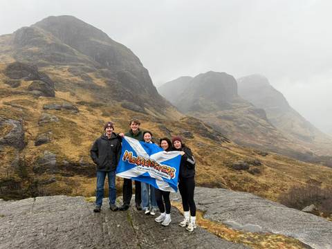       Group with Scottish flag on rocky ledge amid misty Highland scenery
  