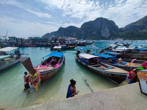       Busy harbour filled with colourful long-tail boats on crystal water against dramatic limestone cliffs.
  