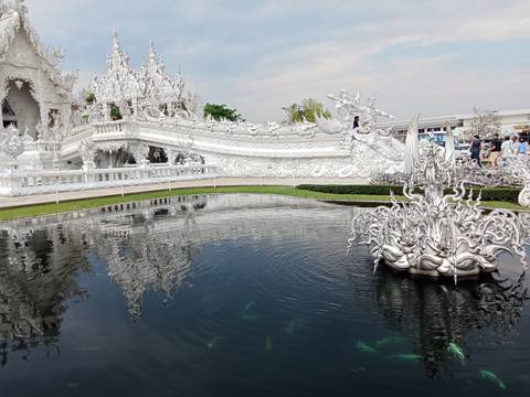       Reflective pool mirrors the ornate white bridge and carvings of Wat Rong Khun while visitors look on.
  
