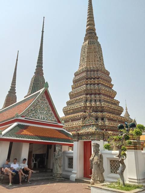       Colorful tiled stupas and a towering prang rise against a pale sky at a Bangkok temple complex.
  