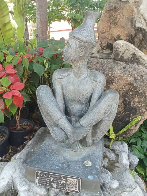       Weathered stone statue of a seated hermit yogi surrounded by tropical plants and flowers.
  