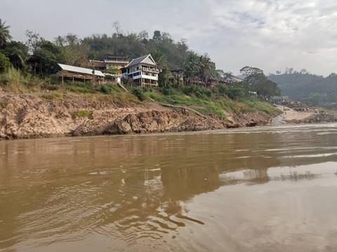       Small riverside village with wooden lodges perched on a rocky bank reflected in the Mekong’s calm water.
  