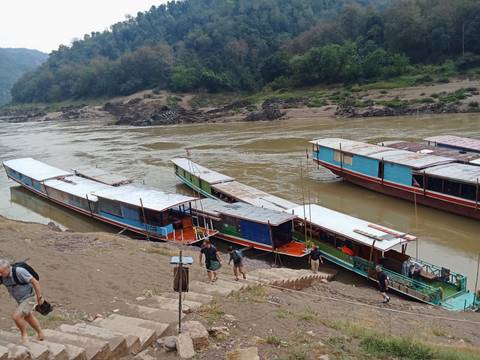       Long narrow boats painted in bright blues and greens are moored on a sandy bank while travellers disembark.
  