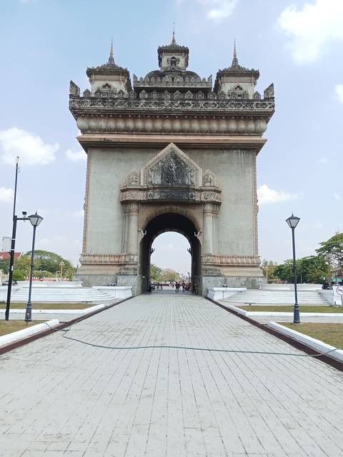       Triumphal arch Patuxai stands tall at the end of a tiled walkway lined with lampposts under a pale sky.
  