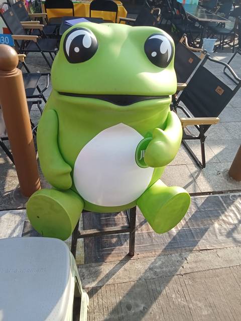       Bright green cartoon frog figurine standing on a pavement in the sun.
  