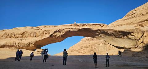       Visitors stand beneath a tall natural sandstone arch set against a deep blue sky in the desert.
  