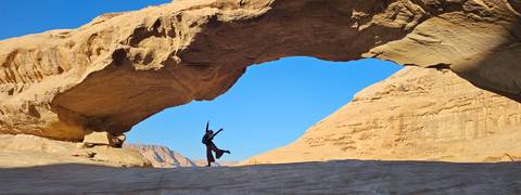       Traveller jumps joyfully beneath a wide natural arch against vivid blue sky and sun-lit desert cliffs.
  