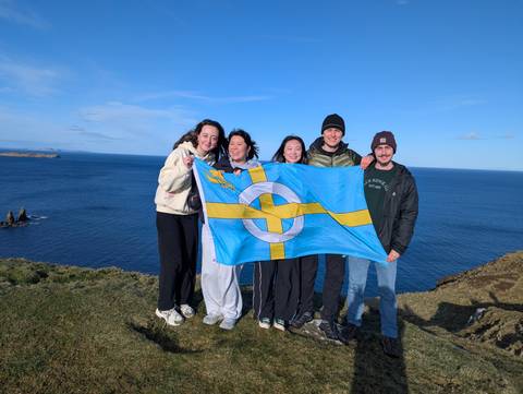       Travellers pose with a blue flag on a windy cliff edge above the vast Atlantic Ocean.
  