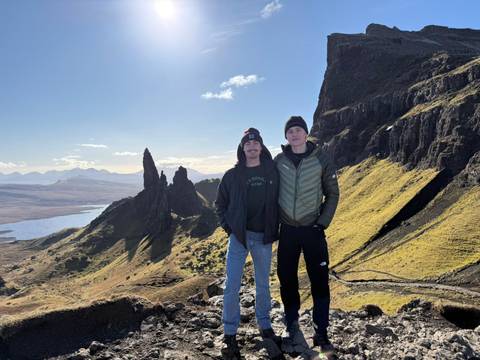       Two hikers stand beneath the jagged rock pinnacles of the Old Man of Storr on a sunny day.
  