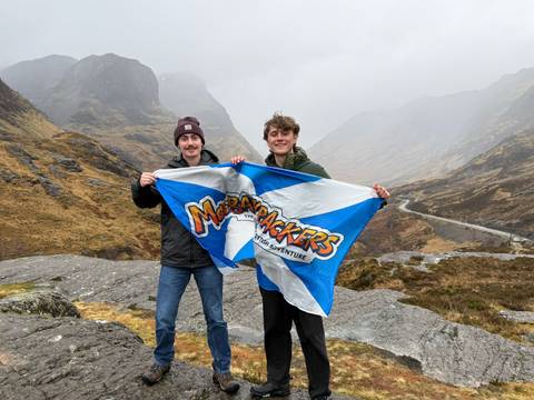       Two friends display a MacBackpackers flag in the dramatic, cloud-shrouded valley of Glencoe.
  