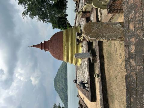       Small brick stupa with golden base by a lake, backed by forested hills and cloudy skies
  