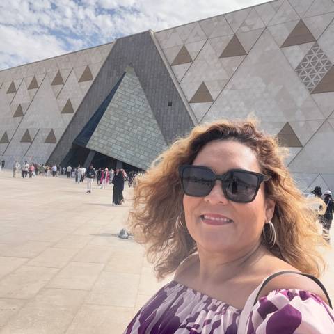       Selfie of a woman wearing sunglasses in front of the geometric facade of the Grand Egyptian Museum with visitors in the plaza.
  