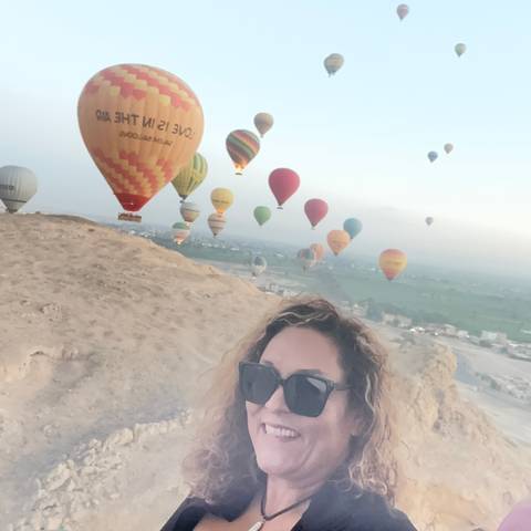       Woman’s selfie with dozens of colorful hot-air balloons rising above desert cliffs at dawn.
  
