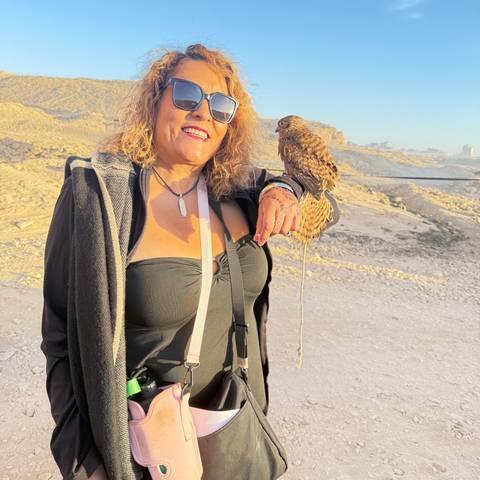       Traveler smiling while holding a small falcon on her gloved hand in a sandy desert area near the pyramids.
  