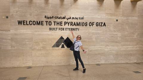       Smiling traveller posing in front of a wall sign that reads 'Welcome to the Pyramids of Giza'
  