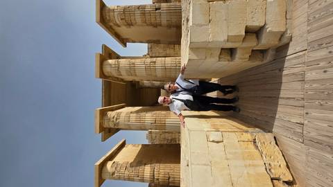       Two travellers stand on a wooden walkway leading to sandy-colored temple columns under a clear sky
  