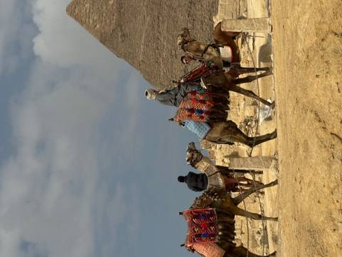       Locals ride decorated camels with a pyramid silhouette against a partly cloudy sky.
  