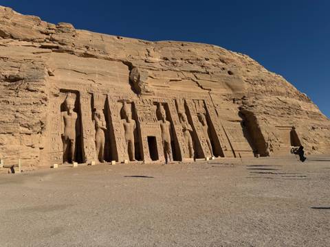       Rock-hewn facade of Abu Simbel’s smaller temple stands under clear sky with wide sandy foreground.
  
