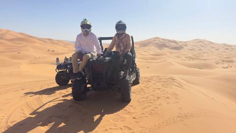       Two riders in helmets sit on an off-road buggy in the middle of sand dunes.
  