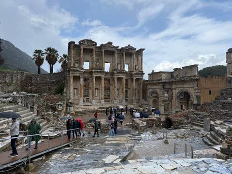       Ancient Roman Library of Celsus facade in Ephesus with tourists holding umbrellas exploring the wet stone ruins on a rainy day.
  