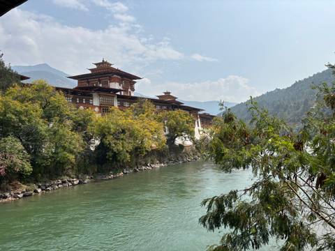       Majestic Punakha Dzong monastery stretches along a jade-green river beneath forested hills and a soft sky.
  
