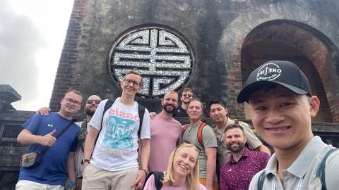       Smiling tour group poses for a selfie in front of an ancient circular mosaic window set into old stone walls.
  