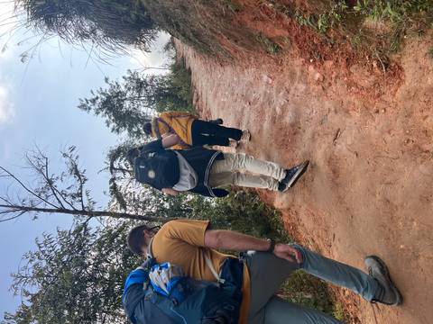       Hikers make their way up a steep reddish dirt trail surrounded by brush and tall grasses.
  