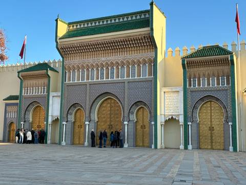       Large ornate golden gates with intricate mosaic tilework at a Moroccan royal palace, with visitors queuing in front under clear blue skies
  