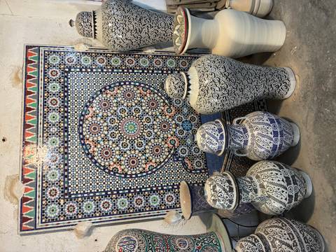       Display of traditional blue-and-white Moroccan pottery in front of a colourful zellige mosaic panel inside a workshop
  