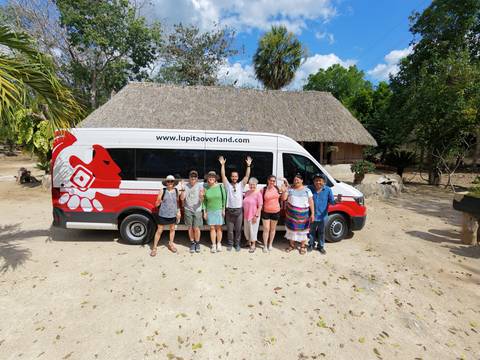       Small tour group cheers beside a branded minibus outside a thatched-roof building on a sunny day.
  