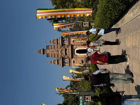       Visitors walk a flag-lined path toward a sandstone Buddhist temple under bright blue sky.
  