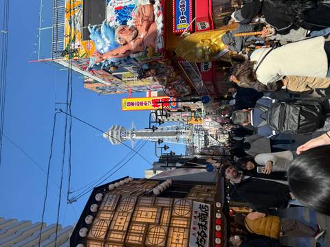       Crowded pedestrian street bursting with colorful shopfronts leading to a towering Tsutenkaku observation tower.
  