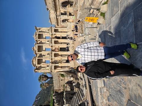       Two tourists pose in front of the grand façade of the Library of Celsus in Ephesus under blue skies.
  